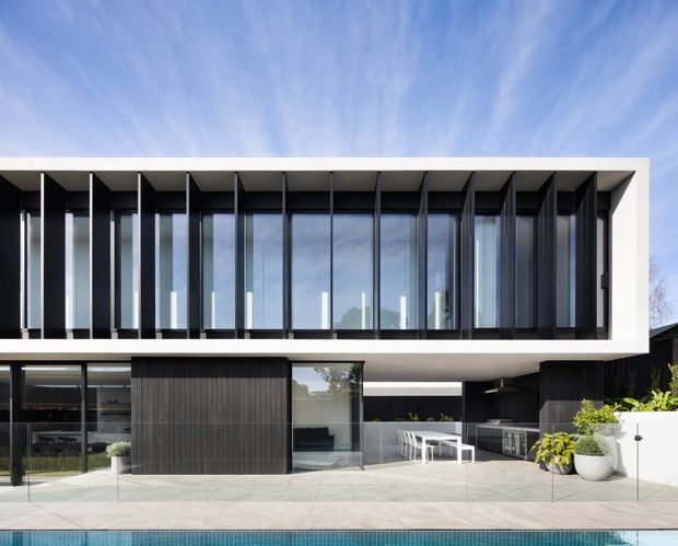 A contemporary two-storey residential facade viewed from the pool terrace. The building features a strong rectangular form, a white projecting frame around a wide upper level of floor-to-ceiling glazing, repeated dark vertical fins, dark timber-look cladding, and a sheltered outdoor entertaining area with dining furniture and built-in cabinetry. A swimming pool occupies the foreground.