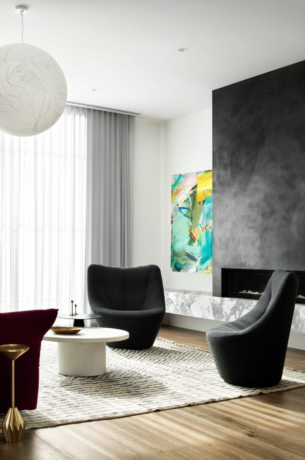 A refined contemporary living room with a bright floor-to-ceiling window dressed in sheer and grey curtains, two sculptural black lounge chairs, a round white coffee table, and a deep burgundy accent chair. A dramatic dark textured feature wall incorporates a linear fireplace above a white marble ledge, while colourful abstract artwork adds contrast. The room has warm timber flooring, a textured neutral rug, and a large spherical pendant light.