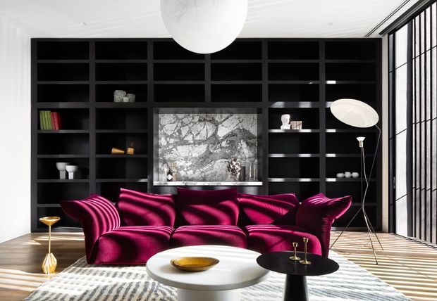 A dramatic contemporary living room with a deep magenta sculptural sectional sofa positioned in front of a full-wall black built-in shelving unit. The shelving incorporates a marble-clad fireplace or feature panel and sparse decorative objects. Floor-to-ceiling black-framed glazing with vertical divisions fills the right side, while a large white globe pendant, sculptural floor lamp, round white coffee table, and patterned rug complete the high-contrast interior.
