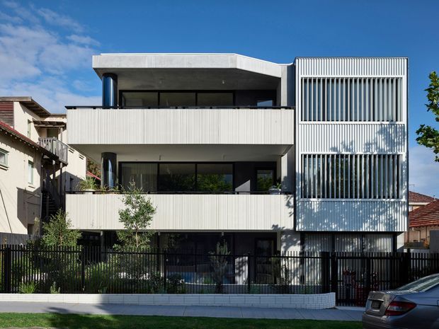 Street-facing view of a contemporary multi-storey residential building with a flat roof, stacked balconies, extensive dark glazing, and prominent white vertical slatted screening on the right-hand facade. The building is set among neighbouring suburban homes with a black metal fence and low rendered boundary wall.