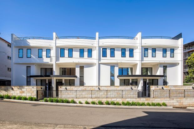 Exterior view of a contemporary multi-unit residential development with three attached white-rendered dwellings. The facade features flat roof terraces with slim metal balustrades, projecting upper-level volumes, broad glazing, recessed balconies, dark entry canopies, and a low rendered masonry boundary wall with metal fencing. The property fronts a paved suburban street under clear blue skies.