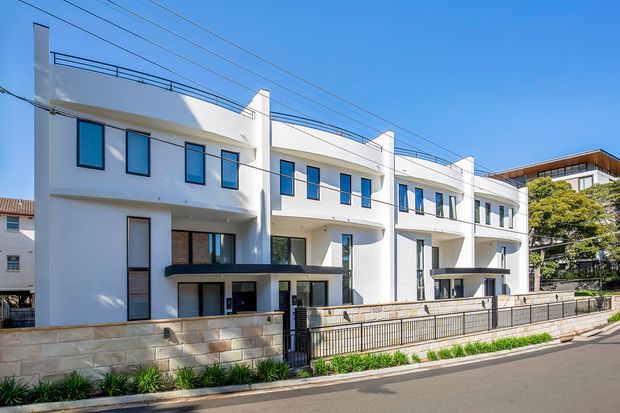 Street-facing view of a contemporary multi-storey residential townhouse development with white rendered facades, curved balcony edges, projecting upper volumes, dark-framed windows, private front terraces, and a low stone-and-metal boundary wall. The building sits in an urban suburban streetscape under a clear blue sky.