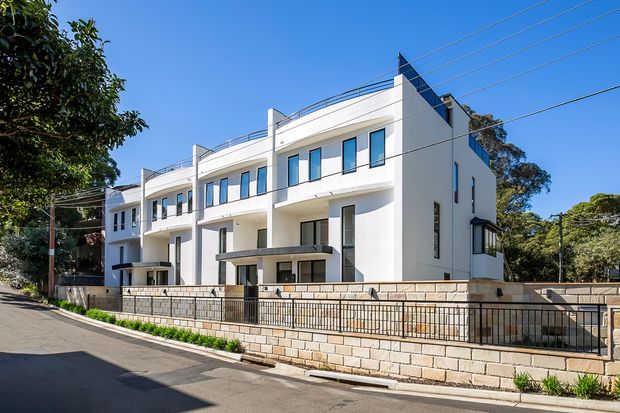 Exterior view of a contemporary multi-storey residential townhouse development with white rendered facades, strong geometric forms, narrow vertical windows, rooftop terraces with metal balustrades, and a covered ground-level entry. The buildings sit behind a low pale stone masonry wall and metal fencing along a suburban street.