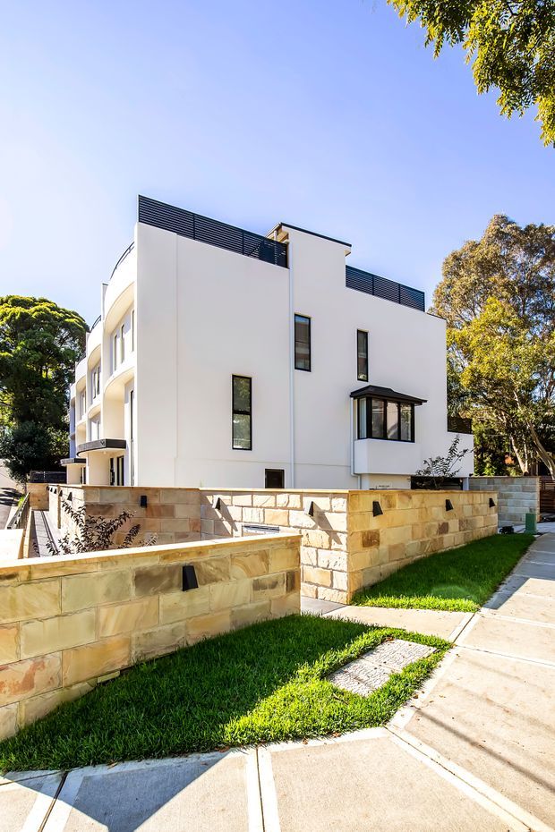 Exterior view of a contemporary multi-storey residential building with smooth white rendered walls, narrow vertical windows, curved balcony edges, dark horizontal rooftop screening, and a projecting glazed bay window. Low warm-toned masonry boundary walls, concrete paving, lawn, and mature trees surround the property.