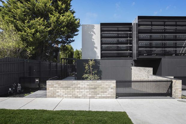 Street-facing exterior view of a contemporary multi-storey residential building. The facade combines dark vertical metal fencing and cladding with projecting horizontal louvre screens, pale rendered walls, and a low light brick masonry planter wall. A concrete driveway and footpath lead to the gated entry, with established trees and lawn in the foreground.