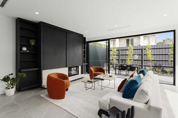 Contemporary living room with a light grey tiled floor, white walls and ceiling, and extensive black built-in joinery incorporating open shelving, concealed storage, and a fireplace or media niche. A large white sectional sofa faces the built-in feature wall, accompanied by rust-orange lounge chairs and slim black coffee tables. Full-height black-framed glazing opens visually to a paved outdoor courtyard with a dark privacy fence and planting.