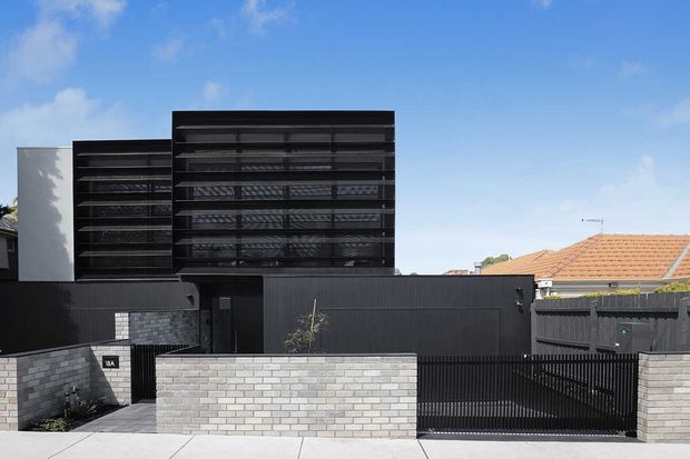 Street-facing exterior of a contemporary multi-level residence with a dark vertically clad lower volume, prominent black louvred upper facade, pale brick boundary walls, and a gated driveway. A neighbouring tiled roof is visible in the background beneath a clear blue sky.
