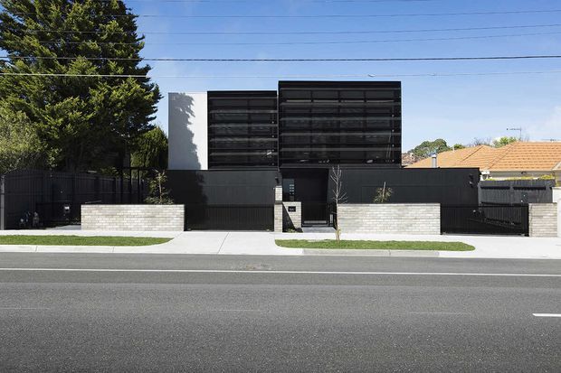 Street-facing view of a contemporary multi-storey residence with a dark rendered facade, broad horizontal screened glazing, a recessed central entry, and pale brick boundary walls. The home is set behind a simple landscaped verge and front fence, with a neighbouring tiled-roof house visible to the side.