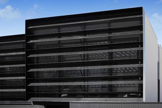Exterior view of a contemporary multi-storey building with a dark facade composed of horizontal louvres or sun-shading screens over glazed levels. A clean white side wall contrasts with the black frontage, and a low metal-clad structure runs along the foreground.
