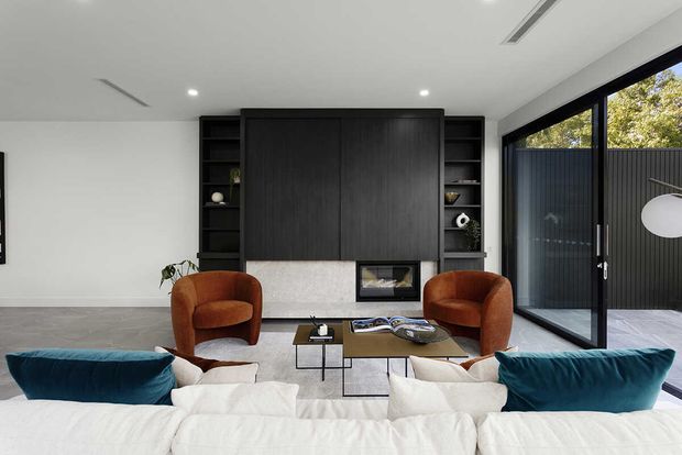 A refined contemporary living room viewed over a large white sectional sofa. The room features a dark timber built-in media wall with open shelving and a low fireplace, a pale stone or concrete hearth surround, two rust-coloured lounge chairs, a slim black coffee table, polished grey flooring, and expansive black-framed glazing opening to a paved outdoor area.