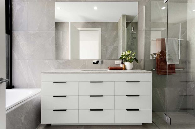 Contemporary bathroom featuring a wide white floating vanity with multiple drawers, a pale stone-look benchtop, large-format light grey wall and floor tiles, a frameless glass shower enclosure, and a large mirrored cabinet or wall mirror. A bathtub is partially visible at left, with black tapware and restrained styling.