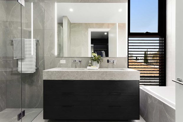Contemporary bathroom with a large double vanity featuring dark textured cabinetry and a pale stone benchtop, a frameless glass shower, wall-mounted mirrors, and a freestanding bathtub beside a slatted black window screen. The room uses large grey stone-look wall and floor tiles with crisp white surfaces and integrated lighting.