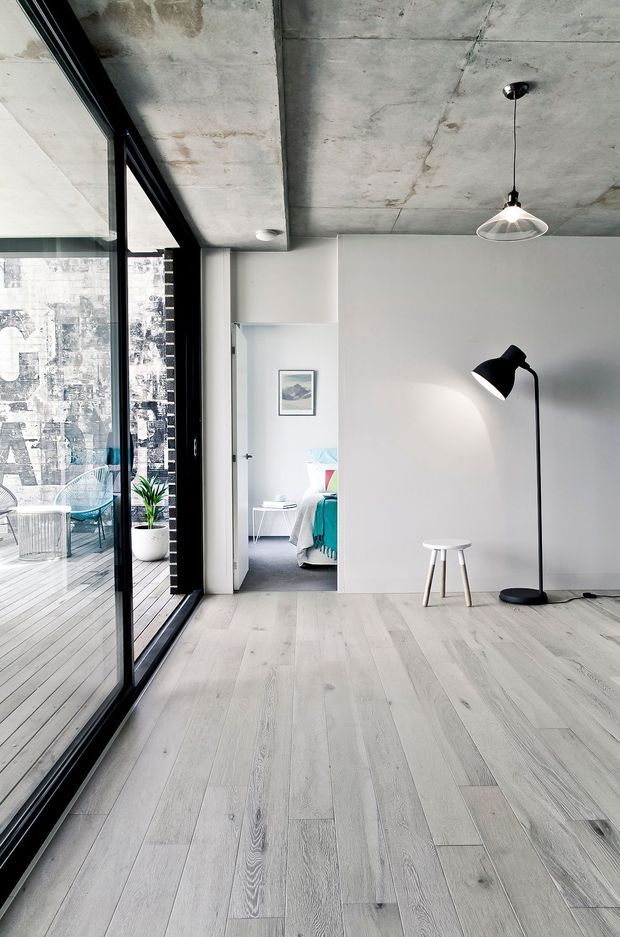 A bright, minimalist interior with pale timber flooring, white walls, and a raw exposed concrete ceiling. Full-height black-framed glazing opens onto a balcony with an urban view. A black floor lamp, pendant light, small stool, plant, and framed artwork furnish the open room, while a bedroom is visible through the doorway.