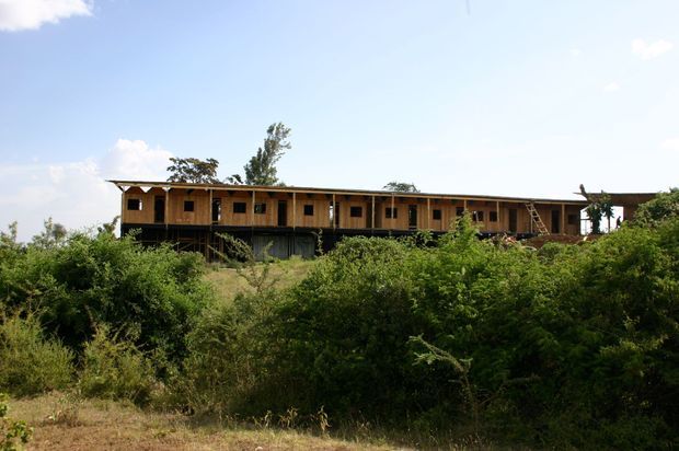 A long, low-rise timber-clad building set within dense natural vegetation. The structure features a simple linear form, a shallow pitched roof with broad eaves, repeated small openings along the upper level, and a darker lower level partially concealed by the landscape. The image is an exterior architectural view in bright daylight.