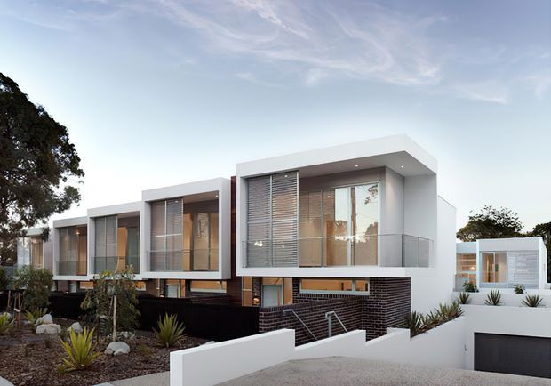 Exterior photograph of a contemporary multi-level residential complex composed of several box-like volumes with white rendered façades, extensive glazing, horizontal external blinds, dark brickwork at the lower levels, and recessed balconies. The property fronts a landscaped garden and driveway, with a separate white garage volume visible on the right. The architecture has strong rectilinear forms and a flat roofline.