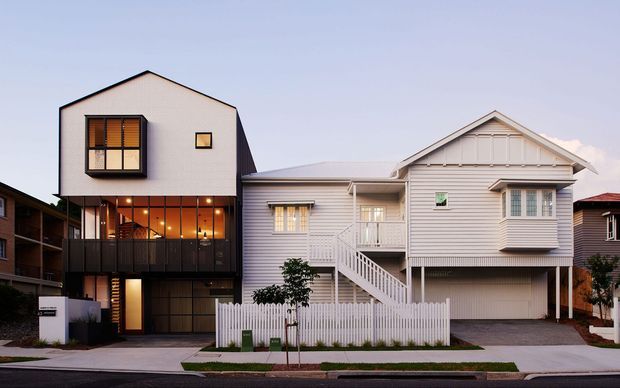 Street-facing view of a compact residential development comprising a contemporary dark-clad multi-storey home beside a light weatherboard gabled house. The homes feature pitched roofs, balconies, upper-level glazing, external stairs, a white picket fence, and an integrated garage, set in a dense suburban streetscape.
