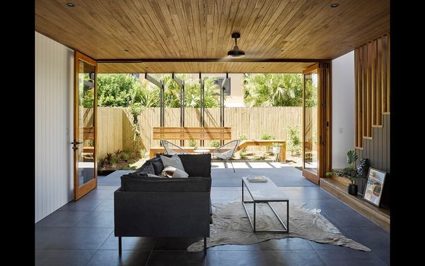 A bright, open-plan living room with large timber-framed bi-fold doors opening onto a paved courtyard. The room features a dark modular sofa, low glass coffee table, concrete-look floor tiles, a timber slatted ceiling, vertical timber wall detailing, and built-in shelving. The courtyard includes timber screening, built-in bench seating, and lush planting.
