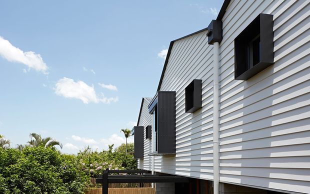 Exterior view of a contemporary multi-level residence with crisp white horizontal weatherboard cladding, projecting dark-framed windows and vertical charcoal feature panels. A timber-toned balcony or deck structure and lush planting are visible below, set against a bright blue sky.