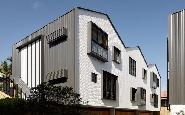 Exterior view of a contemporary multi-storey residential building with a stepped, angular form. The facade combines white textured masonry with dark grey vertically ribbed metal cladding, projecting boxed windows, external louvres, and a partly visible timber-toned lower level. Dense landscaping and neighbouring buildings frame the sunny urban/suburban setting.