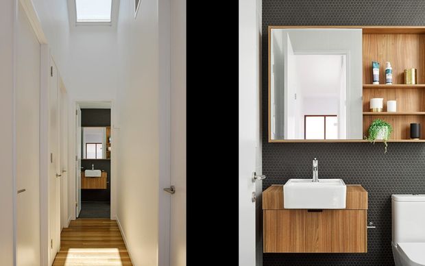 A split-view interior image showing a narrow, bright hallway with white walls, timber flooring, multiple doors and a skylight on the left, alongside a compact contemporary bathroom on the right. The bathroom features dark hexagonal wall tiling, a wall-mounted timber vanity with a white basin, open timber shelving, a large mirror and a partially visible toilet.