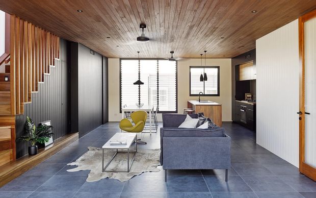 Open-plan contemporary living area with a compact kitchen and dining zone, featuring a timber-lined ceiling, grey tiled flooring, vertical timber stair balustrade, dark cabinetry, a blue-grey sofa, and large windows with blinds. The space has an integrated stairway and a small home office or desk area visible near the rear.