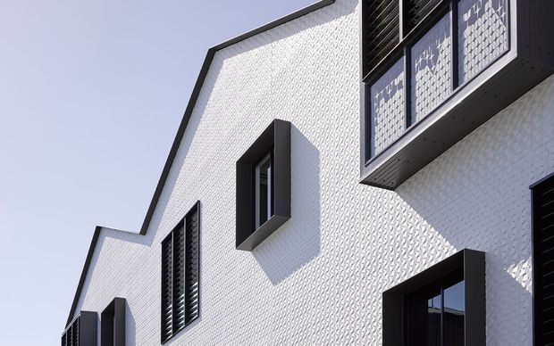 Close-up exterior view of a contemporary multi-level building with white textured masonry or patterned cladding, sharply angled rooflines, projecting dark window surrounds, and black louvred shutters. The facade has a crisp monochrome geometric appearance under bright daylight.