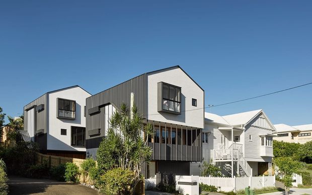 Exterior view of a contemporary multi-level residential development with two prominent gabled volumes, dark vertical corrugated cladding, white rendered walls, projecting box windows, and a screened ground-level balcony. A traditional white weatherboard house sits adjacent, with landscaped planting and a clear blue sky.