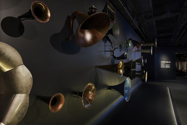 A dark, dramatic exhibition or gallery interior displaying a collection of sculptural metallic horn- and trumpet-like objects mounted across a dark wall. Focused spotlights create strong shadows and reflections on the bronze, brass, and silver-toned surfaces, while a dim corridor and exhibition signage recede into the background.