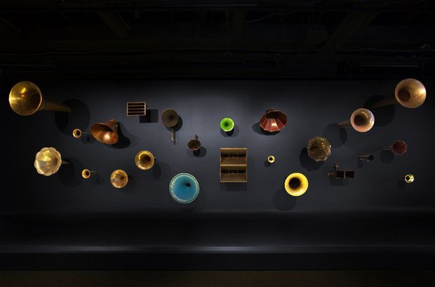 A dark, moody interior installation featuring an eclectic arrangement of sculptural pendant and wall-mounted light fixtures displayed against a charcoal-grey wall. The fixtures vary in metallic finishes and colours, including brass, copper, gold, black, and vivid blue-green, with a central open shelving unit and exposed dark ceiling structure above.
