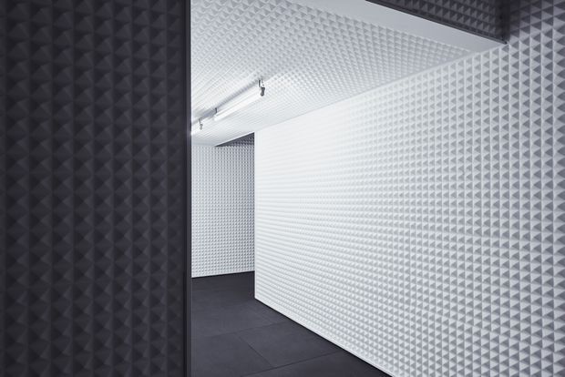 An empty, highly geometric interior corridor or gallery-like space. The walls and ceiling are covered in repeating three-dimensional pyramidal acoustic panels, with a stark white finish contrasted by dark charcoal panels at the left and upper edge. Dark large-format flooring, sharp corners, and minimal track lighting create a monochromatic, immersive, sound-treated environment.