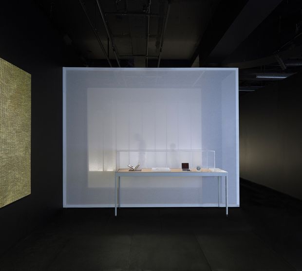 A dark, minimalist exhibition or commercial interior featuring a translucent illuminated white architectural enclosure with a slim pale counter or desk in front. The counter displays a few small objects, while a textured illuminated panel is visible at left. The exposed dark ceiling, black walls, and dark floor create a high-contrast gallery-like setting.
