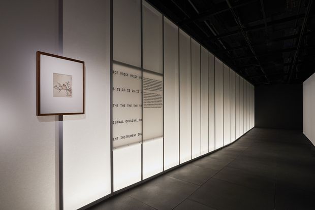 A dark, minimalist exhibition corridor with a long illuminated translucent-panel wall divided by slim vertical frames. A small framed artwork hangs near the entrance, while faint typographic graphics and text appear across the panels. The floor is composed of large dark tiles, and the ceiling is exposed and black, creating a dramatic gallery-like atmosphere.