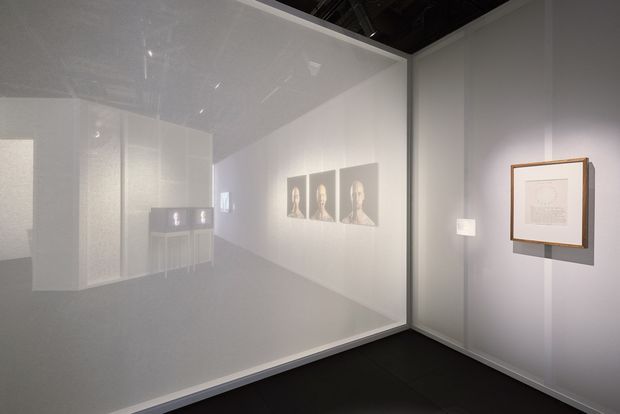 A minimalist gallery or exhibition interior with translucent frosted-glass partition walls, pale grey surfaces, dark flooring, and framed monochromatic artwork displayed on the walls. Reflections and silhouettes of additional artworks and furnishings are visible through the glazing. The space has a restrained, contemporary presentation with subdued lighting.