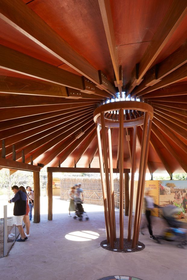 Interior view of a large timber-framed public pavilion or visitor structure. Radiating dark wood beams and slatted wall panels converge around a sculptural central timber column, creating a dramatic vaulted canopy. Open sides lead to an outdoor pedestrian area with informational signage and visitors. The architecture emphasizes exposed structure, warm reddish timber, and a circular central void.