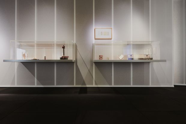 A minimalist interior display wall with two illuminated glass-fronted exhibition cases mounted on a pale grey vertically panelled wall. The cases rest on dark grey shelves and contain small curated objects, with a framed artwork centred above. Dark tiled flooring and controlled gallery-style lighting create a quiet, refined presentation.