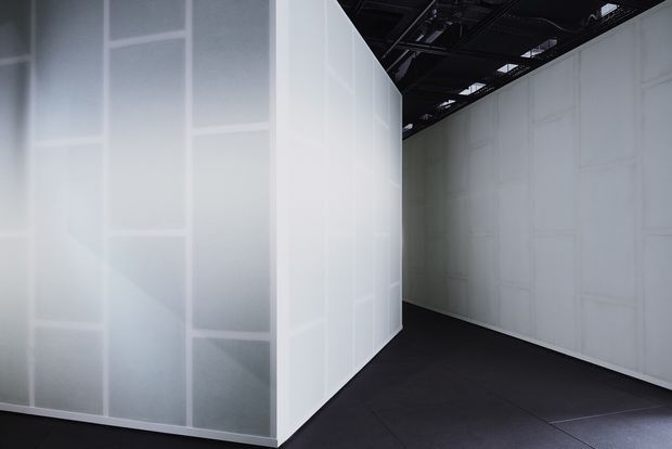 A stark, empty interior space with large translucent white panelled walls forming a corner or corridor. The dark floor contrasts with the pale surfaces, while an exposed black ceiling structure with visible services creates an industrial exhibition or commercial atmosphere.
