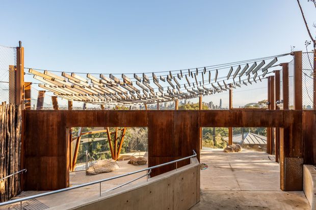 Exterior architectural view of a contemporary steel-and-concrete structure with weathered rust-toned metal walls, exposed overhead framing, chain-link fencing, and a broad concrete circulation area. The open-air space overlooks landscaped surroundings and appears designed as a public or commercial environment.