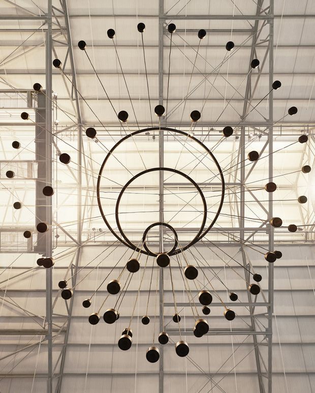 Looking upward into a tall industrial-style interior with an exposed steel structural grid and corrugated metal roof. A dramatic sculptural chandelier composed of concentric dark metal rings and numerous suspended spherical pendant lights dominates the ceiling view. The space is bright, airy, and strongly architectural, with translucent glazing or panels visible behind the structure.