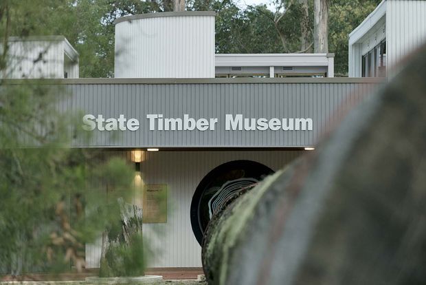 Exterior view of the State Timber Museum, featuring a grey vertically ribbed metal-clad facade, bold raised signage, and a partially visible circular entrance or exhibit opening. The building is set among mature trees and photographed through foreground vegetation, giving the institutional structure a subdued, contextual presentation.