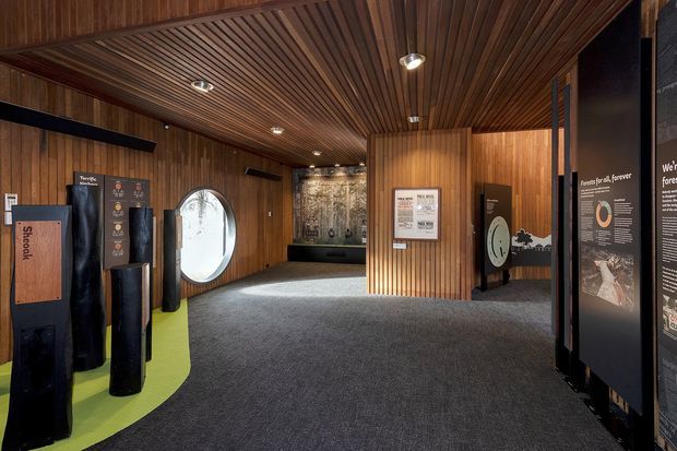 An enclosed exhibition or interpretive space with dark timber wall and ceiling panelling, integrated downlights, charcoal carpet, and freestanding informational display panels. A large oval window and illuminated exhibit graphics are visible, creating a museum or visitor-centre-like interior.