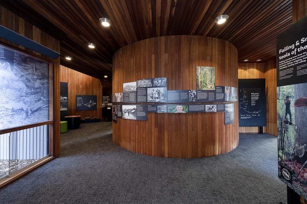 Interior of a museum or exhibition space with a curved cylindrical timber-clad display wall at the centre, surrounded by mounted historical interpretation panels and large graphic displays. The room has a dark timber slatted ceiling with recessed spotlights, dark carpet flooring, and a gallery-like circulation layout.