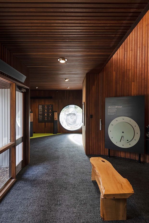 A dimly lit, wood-lined institutional or commercial interior corridor with a richly timber-slatted ceiling and vertical timber wall panelling. Dark carpet runs through the space toward a circular glazed opening, while recessed downlights provide focused illumination. Interpretive display panels and a rough-hewn timber bench occupy the foreground, giving the space the character of a museum, visitor centre, or exhibition building.