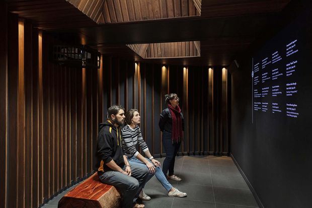 A dark, immersive architectural interior resembling a museum or cultural exhibition space. Visitors sit on a built-in timber bench beside a black wall displaying illuminated text, while vertically slatted dark timber walls, recessed lighting, a timber-lined ceiling, and a skylight-like opening create a dramatic, refined atmosphere.