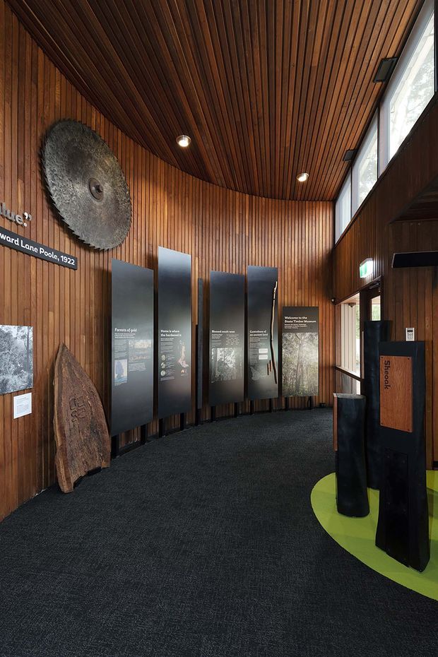 Interior view of a timber-lined museum or visitor-centre exhibition space. The room has a curved vertical timber-slat wall and matching slatted ceiling, dark carpet flooring, tall clerestory glazing, recessed downlights, and freestanding interpretive display panels. Large natural-history objects and signage are integrated into the exhibition, with a visible exit door and reception or information kiosk area.