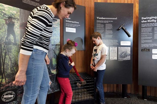 An interactive museum or exhibition display with a timber-panelled interior, dark interpretive information boards, and hands-on educational exhibits. An adult and two children are engaging with a red-handled interactive tool beside the displays.