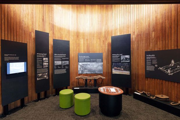 An indoor exhibition or interpretive display space with warm vertical timber wall panelling, dark freestanding informational panels, framed images and text, and small colorful stools around a round display table. The carpeted room has a museum or visitor-centre atmosphere and subdued gallery lighting.
