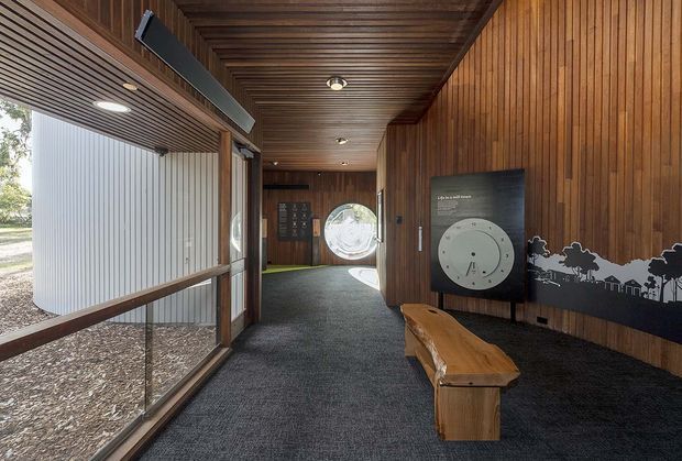 A narrow, museum-like interior corridor with dark timber slatted walls and ceiling, full-height glazing on one side, dark carpet flooring, recessed lighting, and a sculptural solid timber bench. Interpretive display panels and a circular graphic create a gallery or visitor-centre atmosphere, with landscaped grounds visible through the glazing.