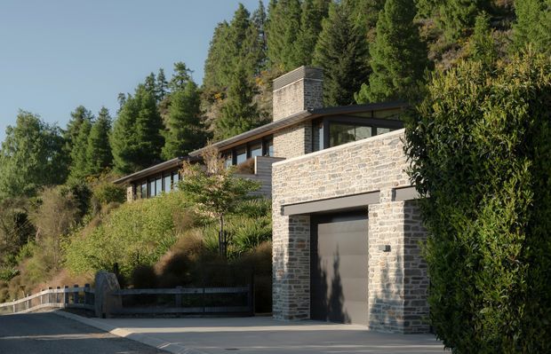 Exterior view of a contemporary hillside residence integrated into a wooded, sloping site. The home features pale stone masonry walls, dark-framed glazing, a low-pitched roofline, a prominent stone chimney, and an integrated garage opening onto a paved driveway. Dense mature trees and landscaped planting surround the building.