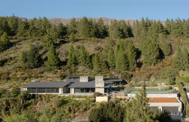 A low, modern single-storey residence integrated into a densely landscaped hillside. The building has dark metal roofing, extensive horizontal glazing, dark cladding, and prominent pale masonry or concrete chimney-like volumes. A swimming pool and adjoining paved terrace are visible beside the house, with rugged, tree-covered hills rising behind it.