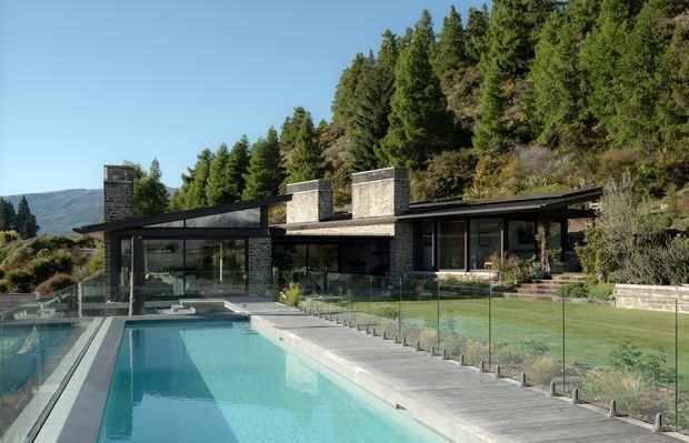 A low, modern single-storey residence set against a wooded hillside, with dark stone and glass walls, broad shallow roof planes, prominent stone chimneys, and extensive glazing. A long rectangular swimming pool with a pale paved deck and frameless glass safety fencing occupies the foreground, while landscaped grounds and a mountain slope sit beyond the house.